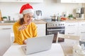 Smiling girl wearing Santa hat having video calling family by webcam. Woman with laptop sitting on kitchen at home using virtual Royalty Free Stock Photo