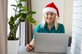 Smiling girl wearing Santa hat having video calling family by webcam. Woman with laptop sitting on kitchen at home using Royalty Free Stock Photo