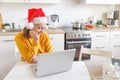 Smiling girl wearing Santa hat having video calling family by webcam. Woman with laptop sitting on kitchen at home using Royalty Free Stock Photo