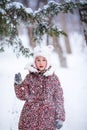 Smiling girl with white fur hat like a cat playing with snow. Winter snowy background and gteen trees