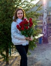 A smiling girl in a white jacket with a bouquet of red roses stands near a Christmas tree. Royalty Free Stock Photo