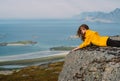 Smiling girl in yellow jacket and sunglasses lies on stone in on the edge of rocks in mountains and looking to fjord Royalty Free Stock Photo