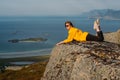 Smiling girl in yellow jacket and sunglasses lies on stone in on the edge of rocks in mountains and looking to fjord Royalty Free Stock Photo