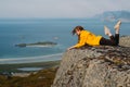 Smiling girl in yellow jacket and sunglasses lies on stone in on the edge of rocks in mountains and looking to fjord Royalty Free Stock Photo