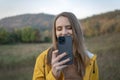 Smiling girl in yellow jacket uses smartphone while walking on autumn day in nature. Young woman photographs beautiful landscape Royalty Free Stock Photo