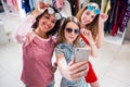 Smiling girlfriends wearing stylish sunglasses having fun time taking selfie with mobile phone while doing shopping in