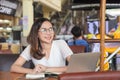 Smiling glasses Asian woman in a white dress sitting in coffee shop, thinking and note to notebook with laptop on table