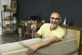 Smiling good-looking brunet hair man in glasses sitting on sofa with victory sign at living room indoor modern flat with Royalty Free Stock Photo