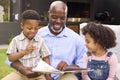 Smiling Grandfather Reading Book With Grandchildren In Garden Together