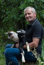 Smiling grey-haired farmer holding two baby goats in his arms.