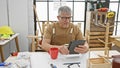 A smiling grey-haired man using a tablet in a carpentry workshop with tools and woodworks visible Royalty Free Stock Photo