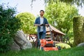 Smiling guy with beard trimming overgrown lawn with electric mower in garden at sunny day. Royalty Free Stock Photo