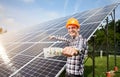 Smiling guy with hundred dollar bill in hand shows at station of solar batteries in which the sun`s rays are reflected
