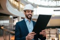 Smiling handsome young arab male engineer, architect or builder with beard in hard hat and suit checks project Royalty Free Stock Photo
