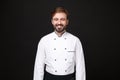 Smiling handsome young bearded male chef cook or baker man in white uniform shirt posing isolated on black wall Royalty Free Stock Photo