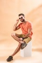 Smiling handsome young man in shorts, summer t-shirt and sunglasses sitting on white cube Royalty Free Stock Photo