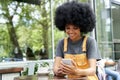 Smiling African hipster teen girl using phone sitting at outdoor cafe table. Royalty Free Stock Photo