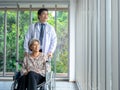 Smiling happy Asian elderly senior female patient sits in wheelchair while man doctor in white coat standing in medical office. Royalty Free Stock Photo