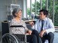 Smiling happy Asian elderly senior female patient sitting in wheelchair talking with friendly man doctor in white coat. Royalty Free Stock Photo