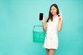 Asian woman holding shopping basket and ok sign and moblie phone on hand isolated on light green background Royalty Free Stock Photo