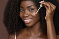 Smiling happy young African black woman applying serum oil on facial skin.