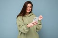 Smiling happy Beautiful young brunette woman wearing stylish green hoodie using mobile phone writing sms isolated on Royalty Free Stock Photo