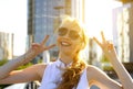 Smiling happy blond girl showing double v-sign standing in city street under sunbeams Royalty Free Stock Photo