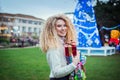 Smiling happy blonde curly woman with tower of colorful gift boxes in hands, enjoying time outdoors in decorated park with