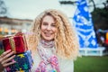 Smiling happy blonde curly woman with tower of colorful gift boxes in hands, enjoying time outdoors in decorated park with