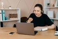 Smiling happy businesswoman in headphones sits at desk, looks at laptop screen, making notes, participating Royalty Free Stock Photo