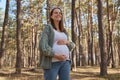 Smiling happy delighted Caucasian pregnant woman waking in spring forest wearing casual clothing rouching her big belly looking Royalty Free Stock Photo