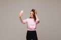 A smiling happy girl in a pink blouse takes a selfie on a smartphone on a gray background