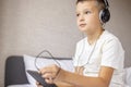 Smiling happy kid caucasian boy in white T-shirt siting on bed, using tablet computer with headphones in bed at home Royalty Free Stock Photo