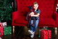 Smiling happy little boy sitting and holding teddy bear on the red coach near the chrismas tree Royalty Free Stock Photo