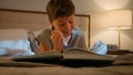 Smiling happy little cute caucasian child kid boy schoolboy reading interesting book evening in bedroom holding Royalty Free Stock Photo