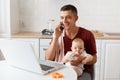 Smiling happy man wearing burgundy casual t shirt with towel on his shoulder, looking after baby and working online from home, Royalty Free Stock Photo