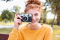 Smiling happy red hair student girl taking picture with old vintage camera outside in autumn park