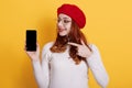 Smiling happy red haired female holding mobile phone with blank screen, pointing at device with fore finger, girl curls standing Royalty Free Stock Photo