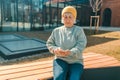 Photo portrait of blonde happy woman 60s holding phone sitting on bench in city spring park outdoors resting use mobile Royalty Free Stock Photo