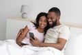 Smiling happy young african american boyfriend and girlfriend hugging, sitting on bed in bedroom interior Royalty Free Stock Photo