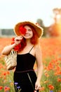 Smiling and happy young red-haired  woman in a black dress and a hat holding red poppies near the face. Beautiful poppy field at Royalty Free Stock Photo