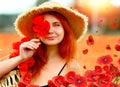 Smiling and happy young red-haired woman in a black dress and a hat holding red poppies near the face. Beautiful poppy field at
