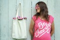 Smiling hippie woman in pink t-shirt with Be Cheerful inscription and pink round glasses standing near blank canvas eco shopping