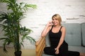 Cheerful blonde woman talking on mobile phone while sitting on comfortable couch near green plant. Royalty Free Stock Photo