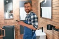 Smiling, holding smartphone in hands. Plumber in blue uniform is at work in the bathroom