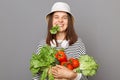 Smiling hungry Caucasian woman wearing striped shirt and panama holding organic vegetables standing isolated over gray background