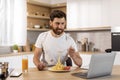 Smiling hungry handsome adult caucasian man in white t-shirt watching video on laptop, eating pasta