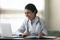 Smiling Indian female doctor physician using laptop, taking notes