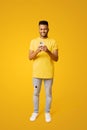 Smiling indian man using smartphone sitting on the floor in relaxed pose, isolated on yellow backdrop, carefree serene Royalty Free Stock Photo