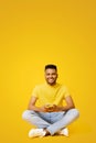 Smiling indian man using smartphone sitting on the floor in relaxed pose, isolated on yellow backdrop Royalty Free Stock Photo